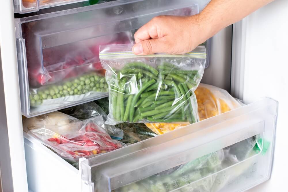 A close up of a hand placing a sealed bag of green beans into a produce drawer with other vegetables.