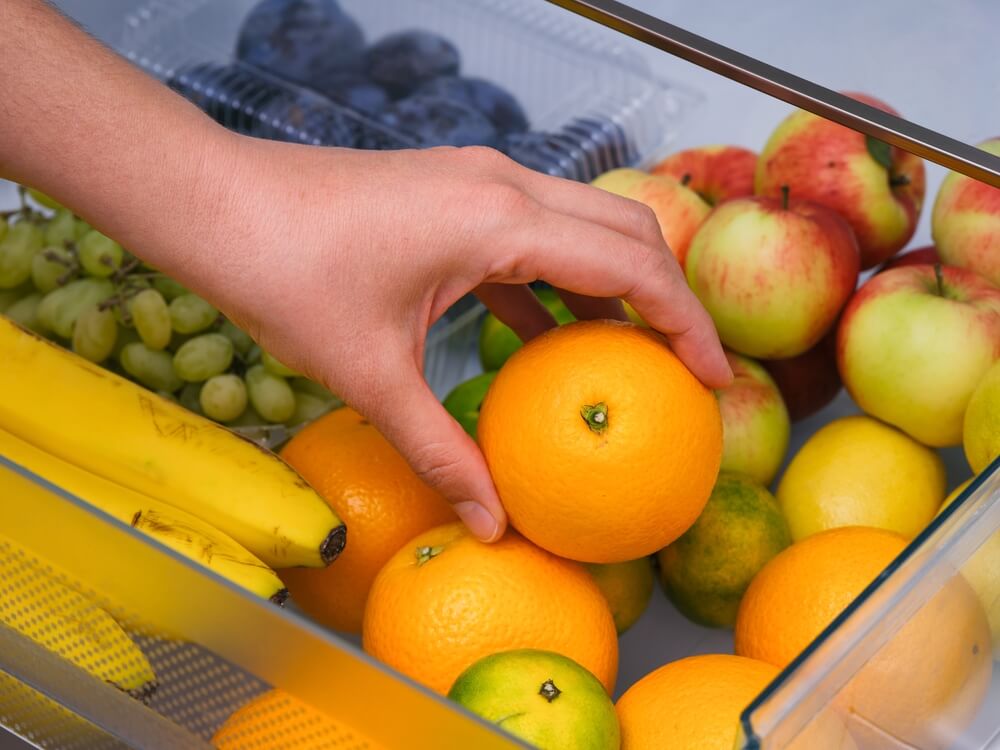 A close up of a person's hand placing an orange in a produce drawer of a stocked fridge.