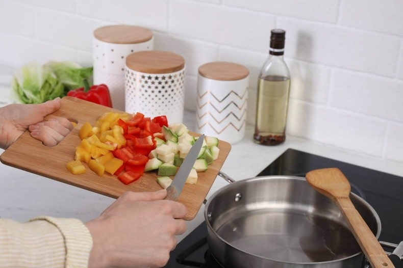 two hands pushing chopped vegetables from a cutting board into a pan on the stovetop