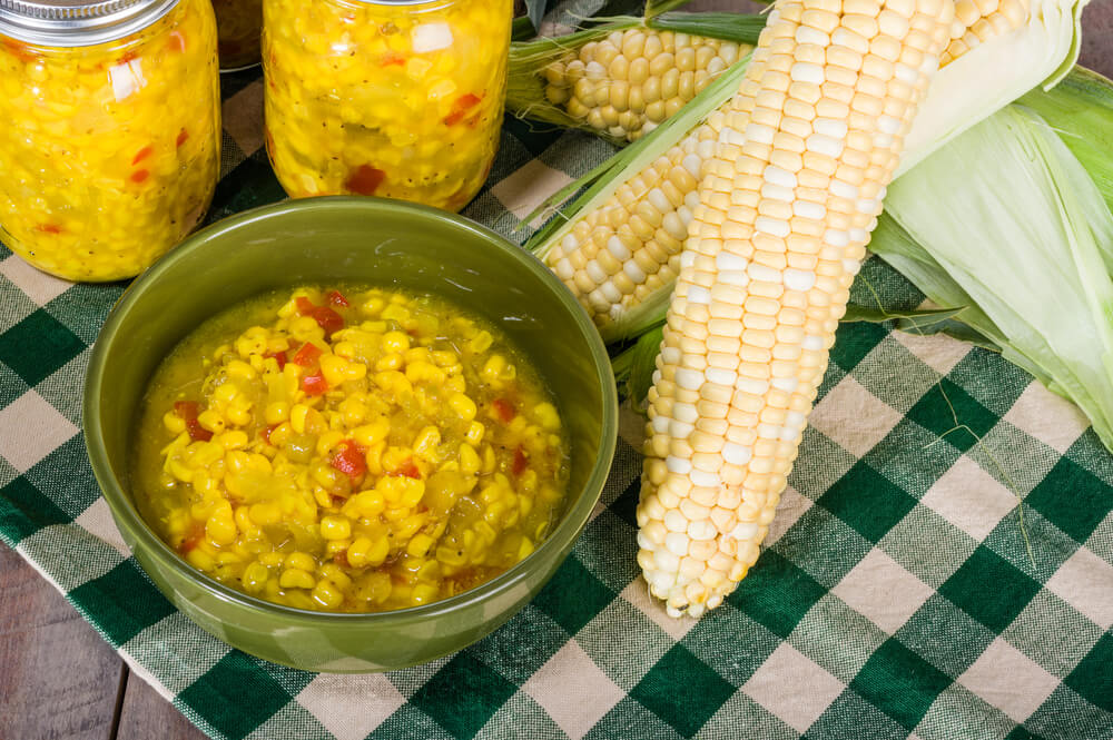 a bowl of pa dutch corn relish sitting in front of 2 cans and 2 ears of corn